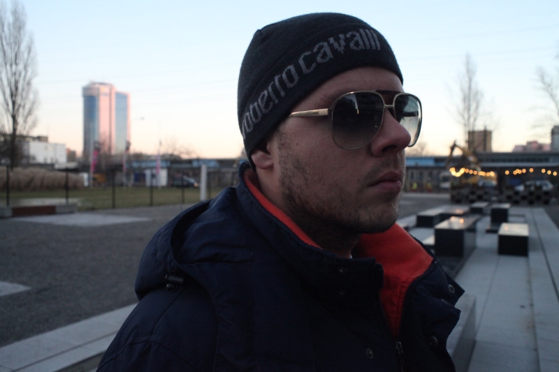 Na zdjęciu widnieje raper Kaz Bałagane w jesiennej czapce, okularach przeciwsłonecznych i kurtce, a w tle widoczny jest rozmazany wiadukt.